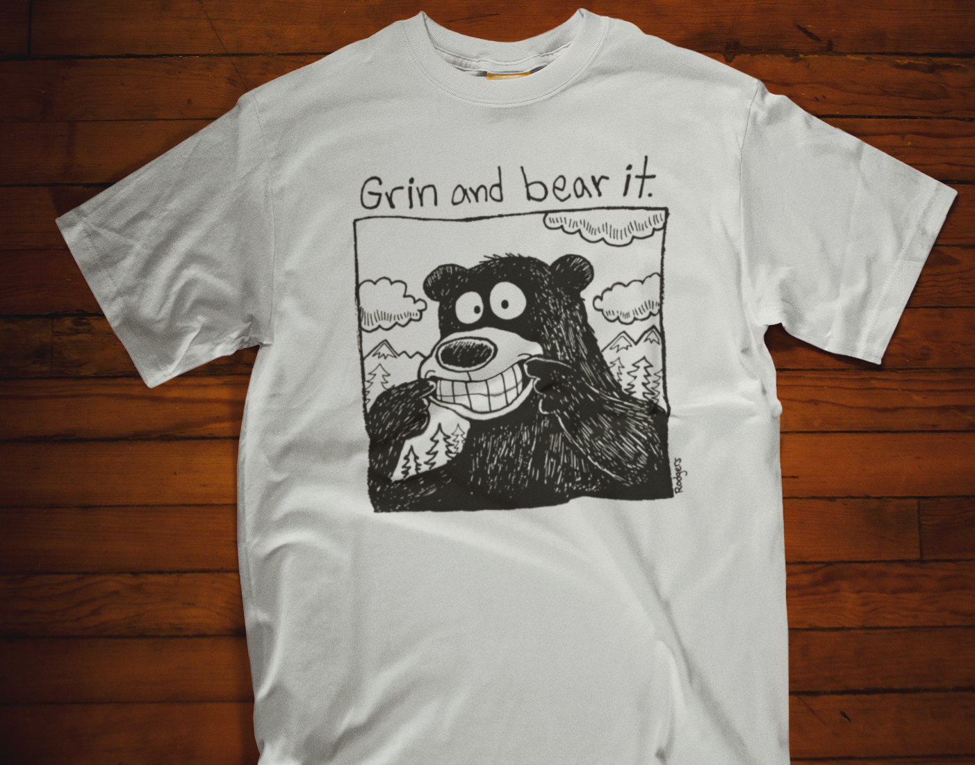 White t-shirt with a bear graphic and text 'Grin and bear it' on a wooden surface
